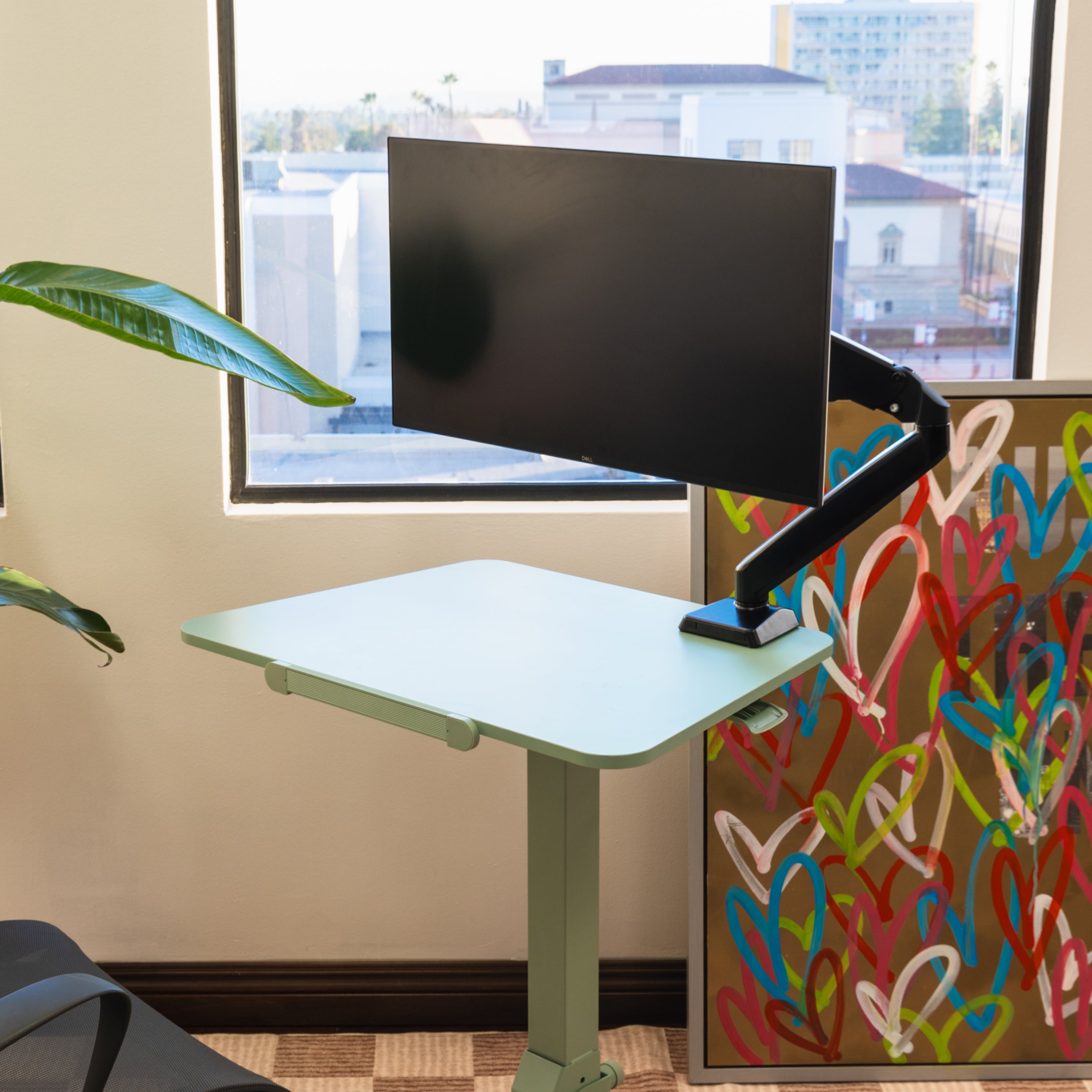 Adjustable standing desk with a monitor in a room with a window and colorful wall art.