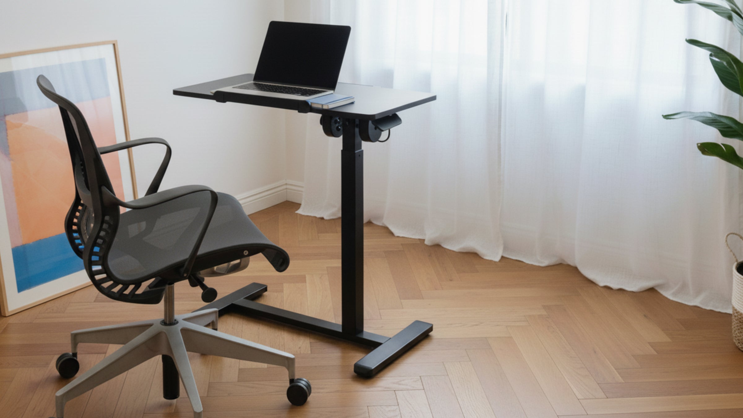 Black adjustable desk with a laptop on a wooden floor in a room with white curtains and a plant.