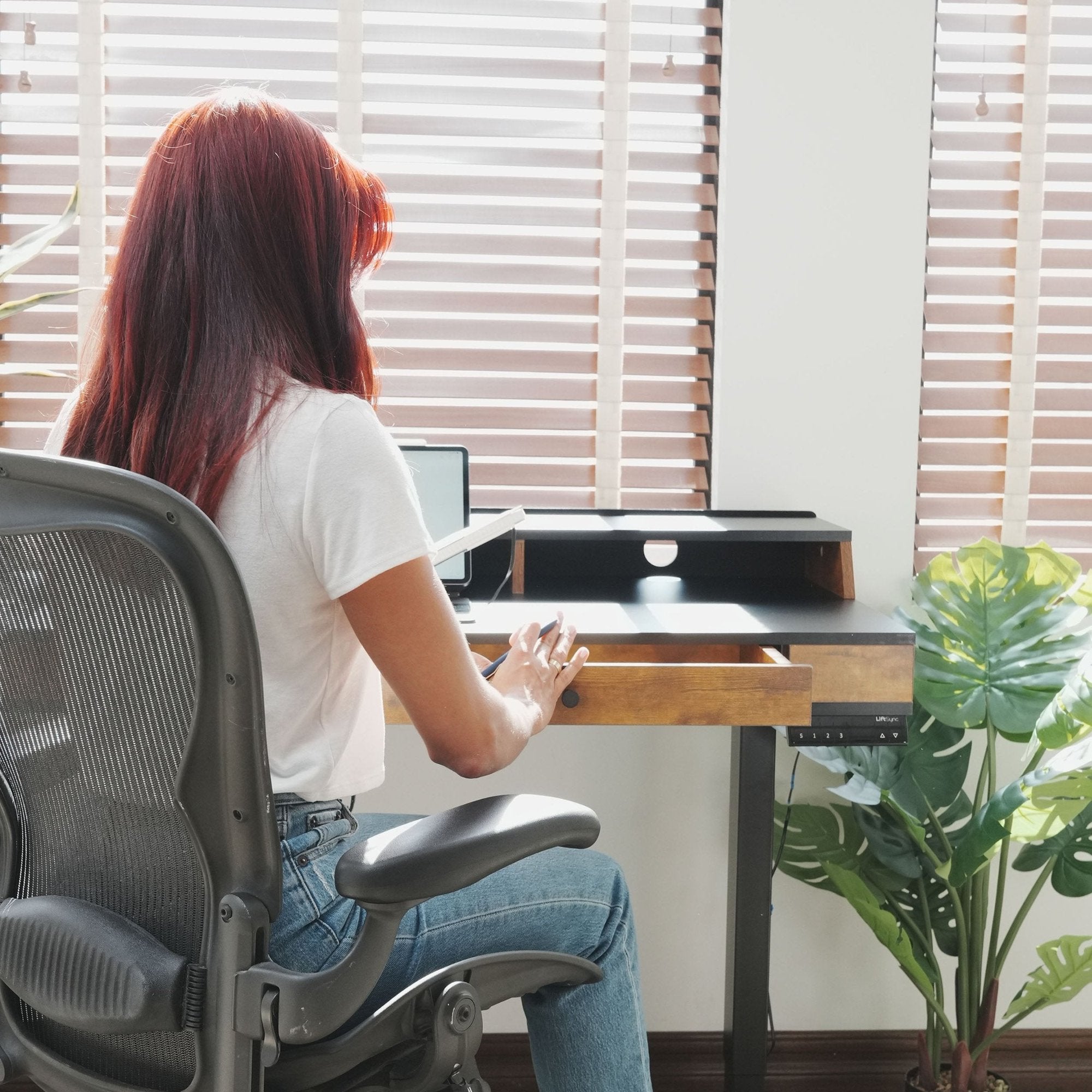 Woman sitting at LiftSync Studio standing desk with integrated shelf and storage drawer