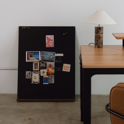 Black magnetic board with various small posters on a white wall next to a wooden table.