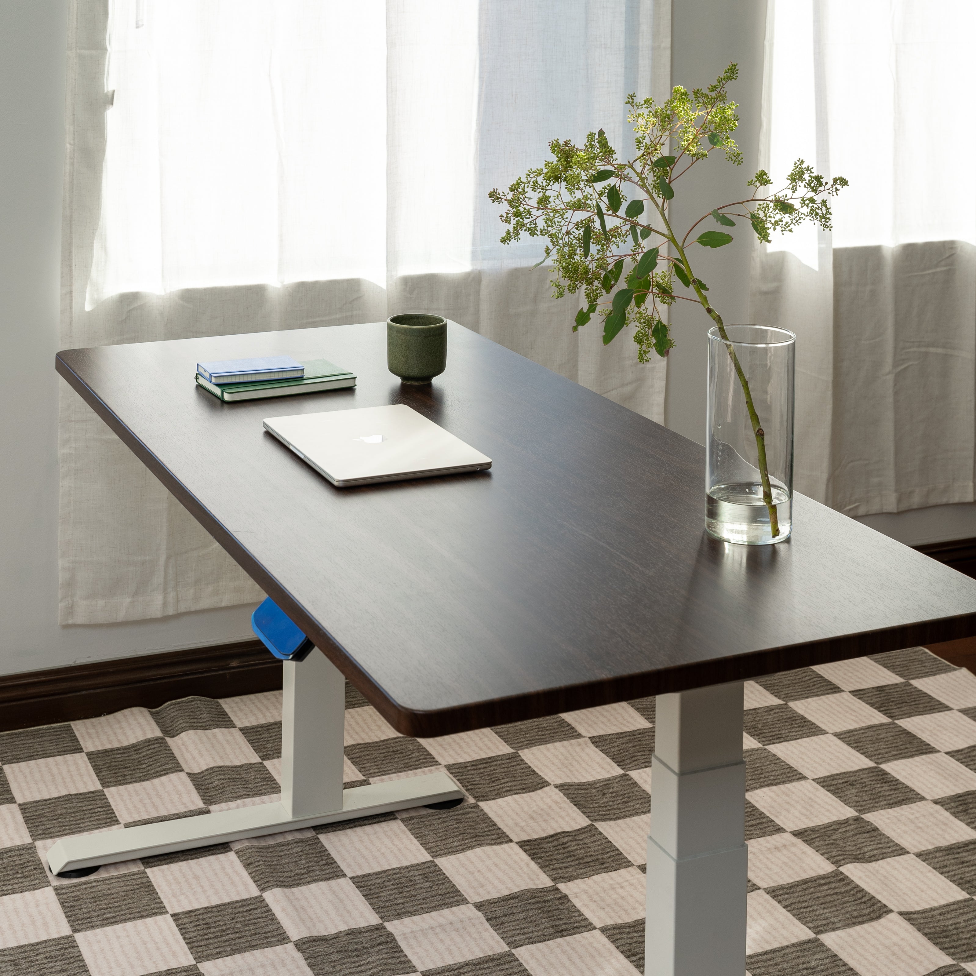 Adjustable desk with a laptop, books, and a vase on a checkered floor.