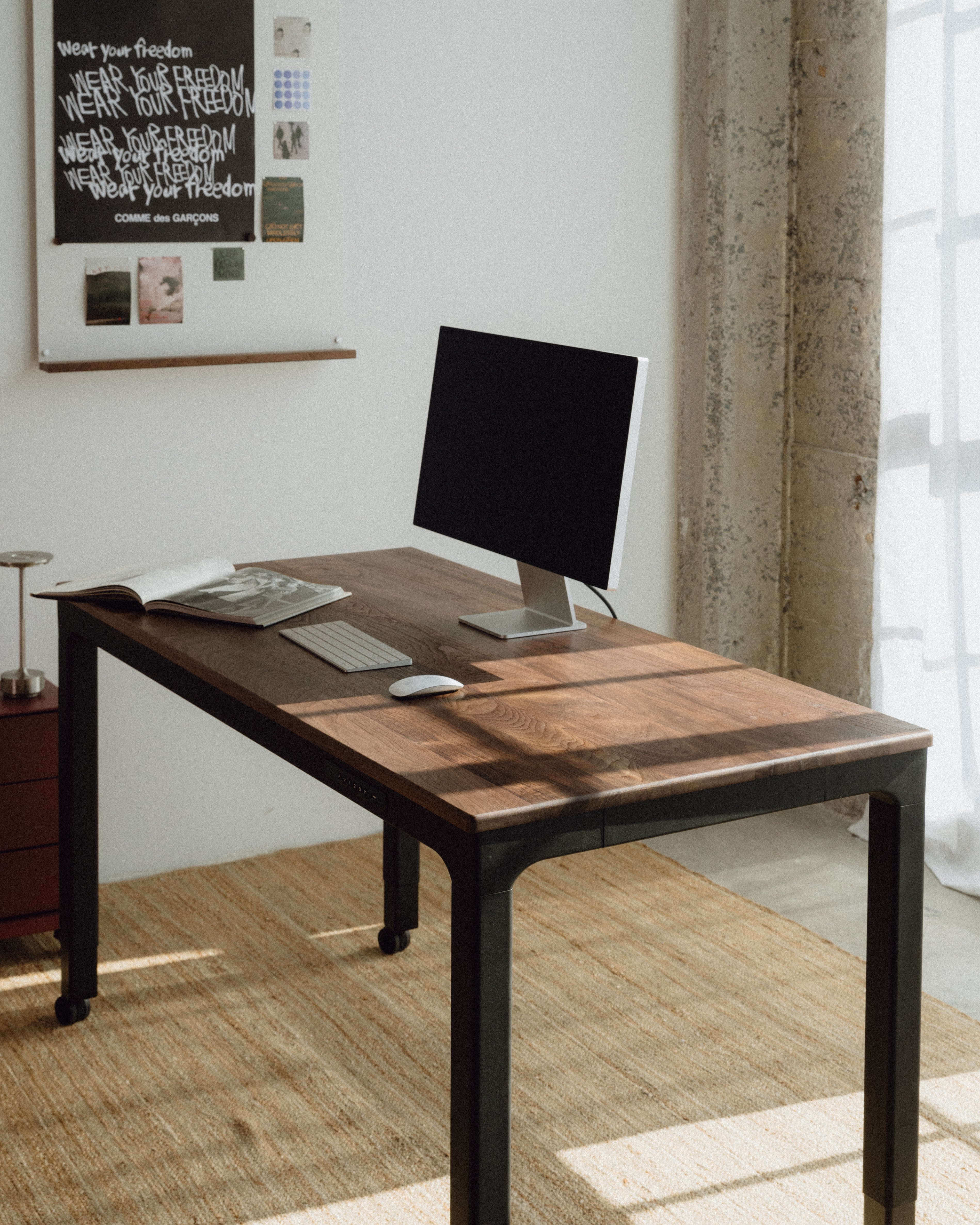 walnut desktop standing desk with computer setup in a room with a window and wall decorations.